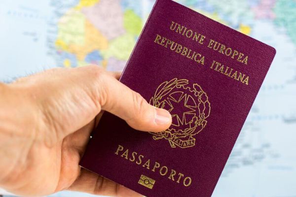 una mano di un uomo che tiene un passaporto italiano. Sullo sfondo, una cartina geografica